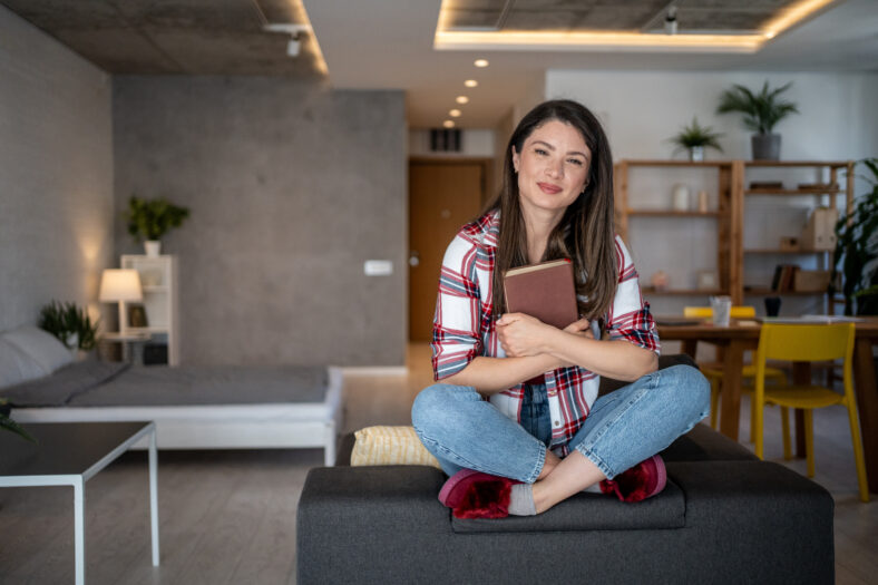 Young woman sitting cross-legged on a sofa, embracing a book in her modern, cozy apartment, savoring a tranquil moment of leisure and comfort in her domestic space