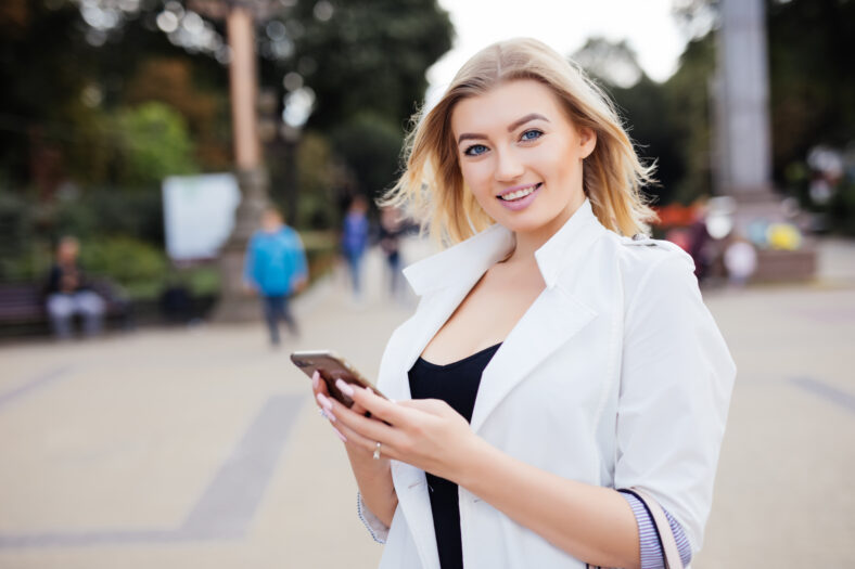 gorgeous beautiful young woman with blonde hair messaging on the smart-phone at the city street background. pretty girl having smart phone conversation in sun flare.