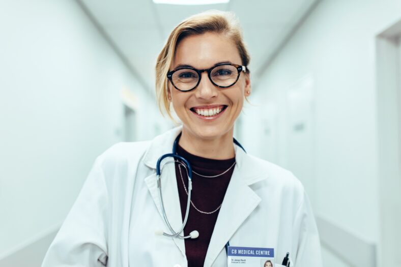 Portrait of happy young female doctor standing in hospital corridor. Caucasian woman working in healthcare center.