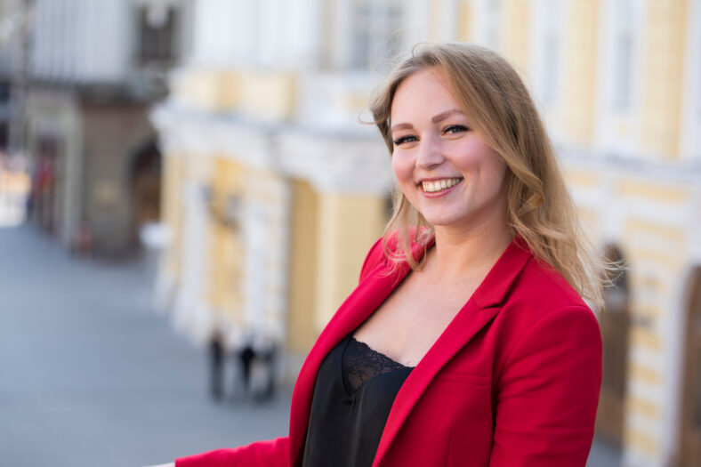 Portrait close up of young beautiful happy blonde woman in red jacket, summer street outdoors