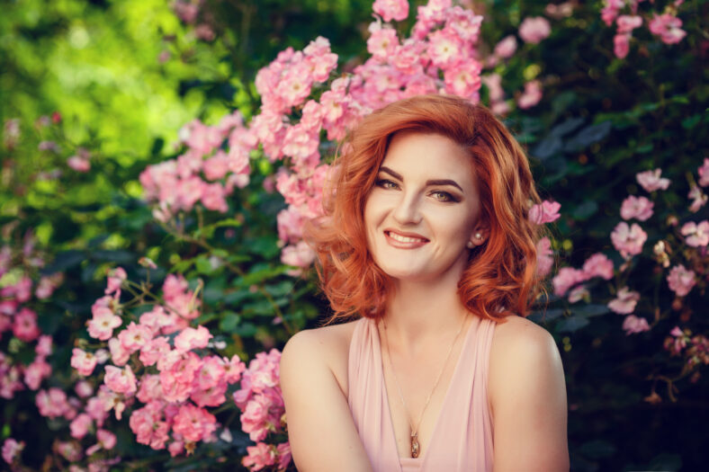 Lovely young woman in a pink dress and red hair, waiting for a date. A walk through the summer city, pink flowering bushes