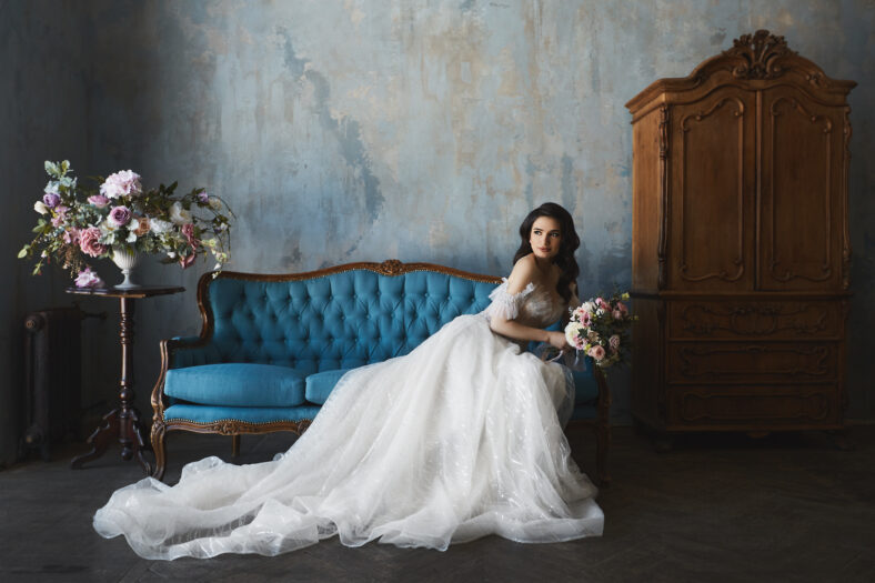 beautiful brunette model girl in stylish and fashionable lace wedding dress with naked shoulders sits on the antique sofa and posing in luxury vintage interior.