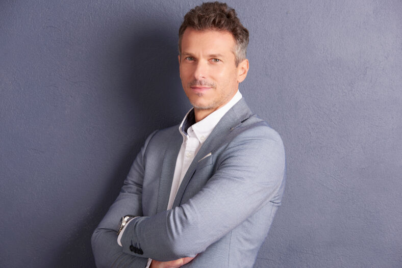 Close-up portrait shot of confident middle aged businessman with folded arms standing at grey wall and looking at camera.