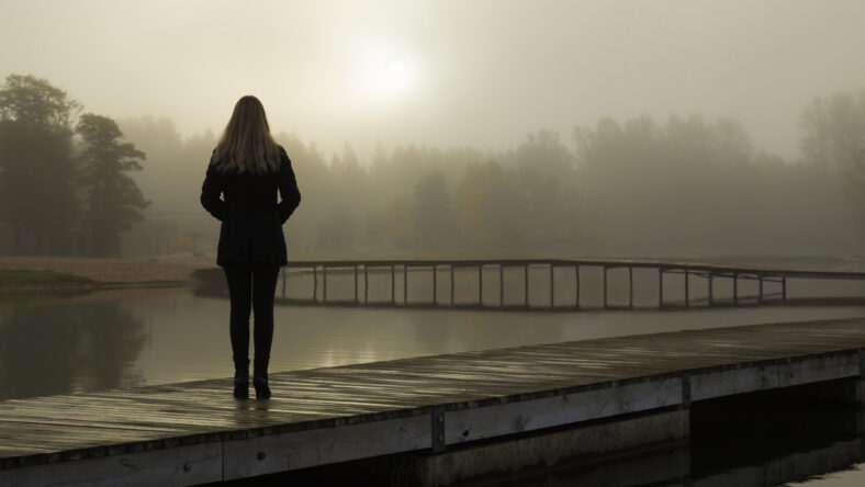 Young woman standing alone on lake footbridge and staring at sunrise in gray, cloudy sky. Mist over water. Foggy air. Early chilly morning. Dark, scary moment and gloomy atmosphere. Back view.