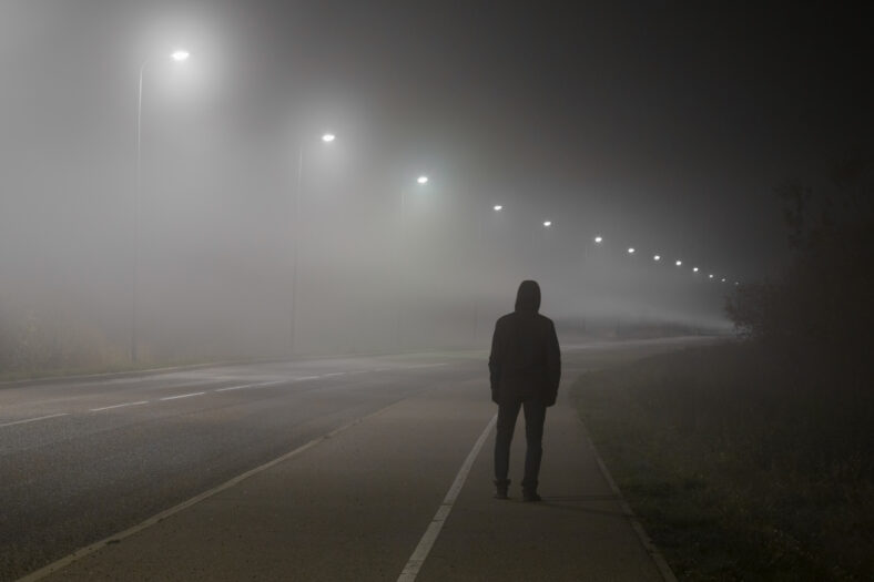 Young man alone slowly walking under white street lights in night. Dark time. Peaceful atmosphere in mist. Foggy air. Back view.