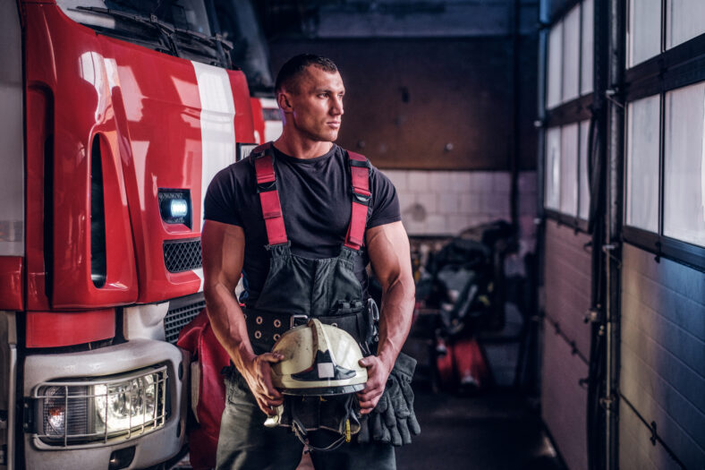 Muscular fireman holding a protective helmet in a garage of a fire department, standing next to a fire engine and looking outside