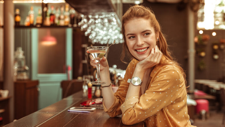 Holding a drink. Smiling glowing happy contended red-haired appealing bewitching woman holding a martini drink in a hand sitting at the bar counter.