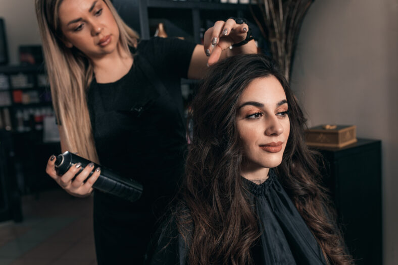 Female hairdresser makes hairstyle on young woman with brunette hair in salon.