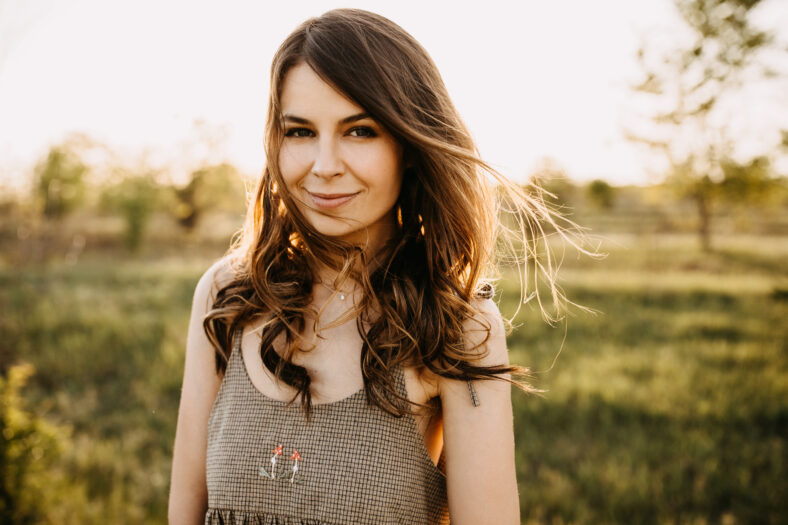 Close-up portrait of a young brunette woman with long wavy hair, standing in a field, outdoors, smiling, on summer day.