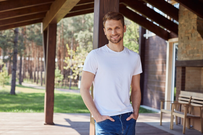 Handsome pleased male standing near his house with hands in pockets