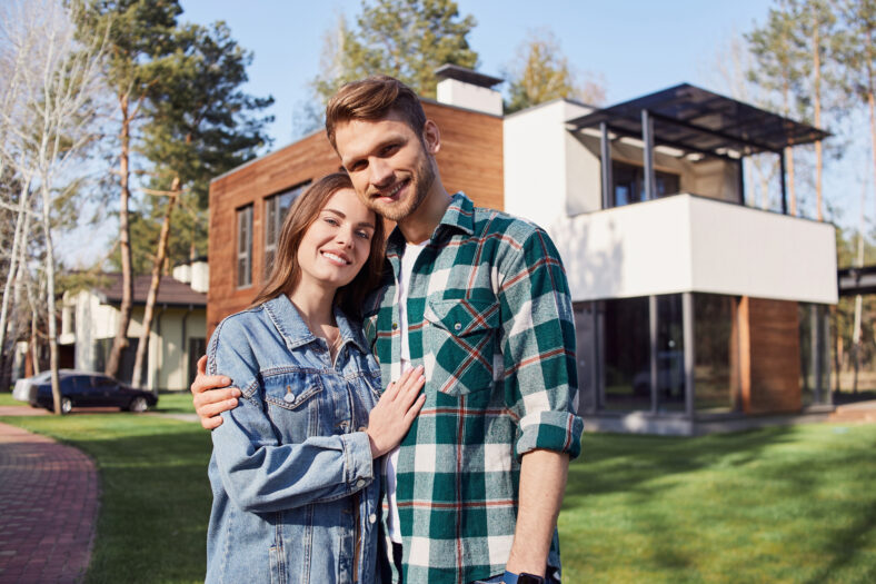 Waist up of smiling loving couple posing outdoors with their new house on the background