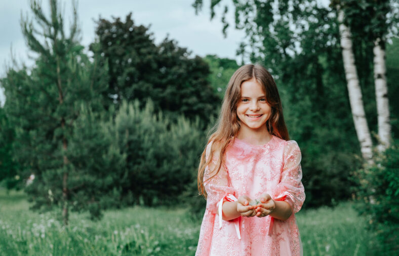 teenage girl with long hair in a pink dress smiles against a background of greenery