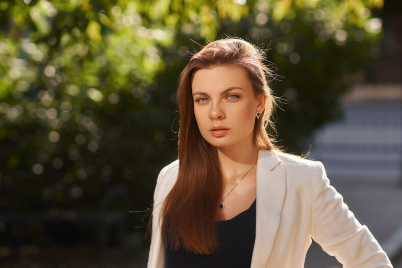 Young woman in black shirt, jeans and white blazer walking and posing outdoors on a summer day. Sun backlight. Outdoor portrait