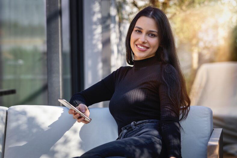 Portrait of a young woman with long, black hair sitting on a terrace and holding her mobile phone