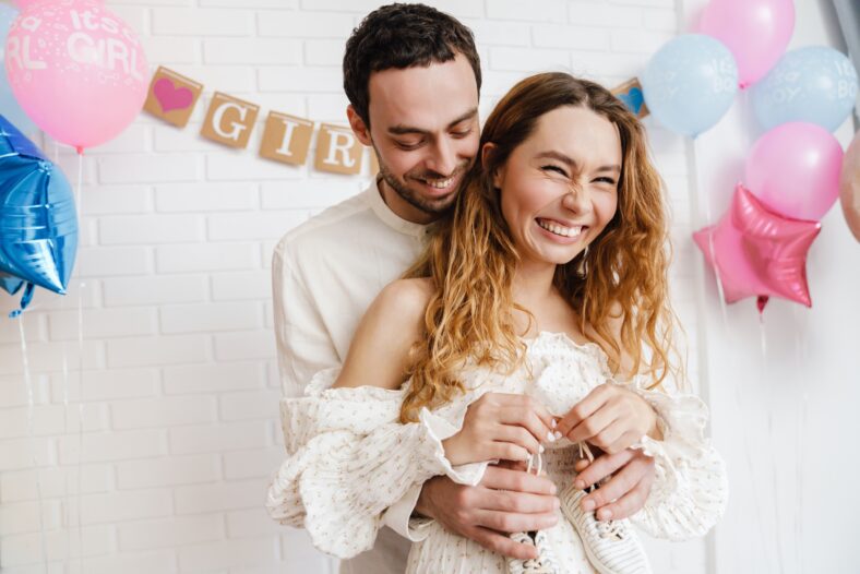 Young happy couple posing with baby shoes during gender reveal party indoors