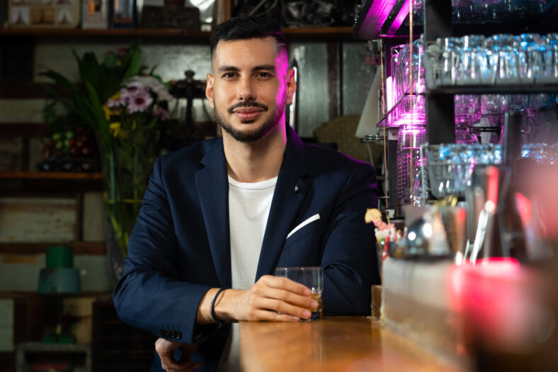 Handsome Caucasian man sitting at bar counter holding whiskey glass with ice cube and drinking alcoholic drink from barman in nightclub. Nightlife holiday party celebration and small business concept