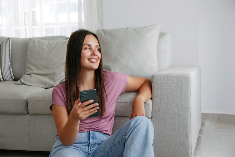 Portrait of young brunette woman wearing mom jeans sitting on the floor leaning on the couch texting and smiling. Joyful female having fun video phone call. Background, copy space, close up.