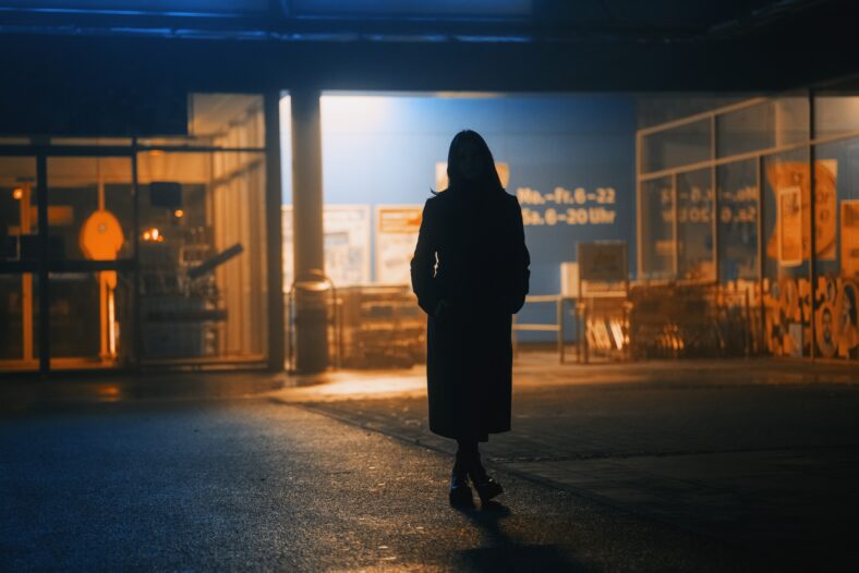 dark silhouette of a girl dressed in a long coat against the background of a night city with advertising showcases