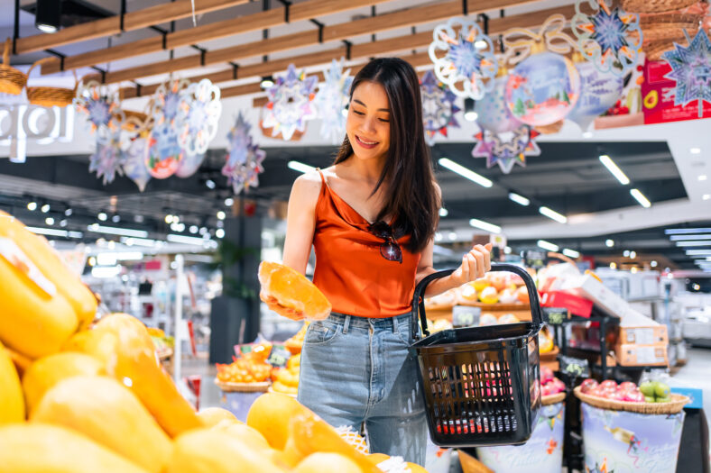 Asian young beautiful woman holding grocery basket walk in supermarket. Attractive female holding vegetable and fruits picking up from shelf for health and wellness in shopping department center.