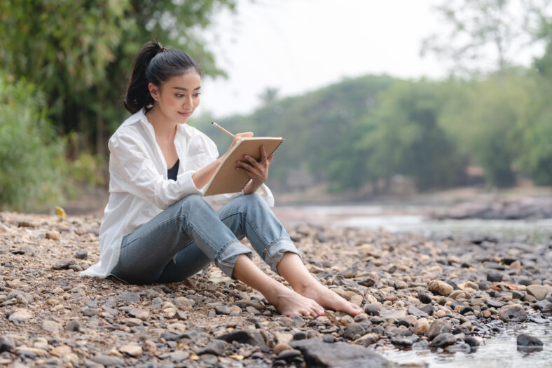 Photo of young attractive Asian woman writing on diary while sitting outdoors in the park, Writer sitting in garden, Female freelance are relaxing to writing story on jobs, beautiful Asian women