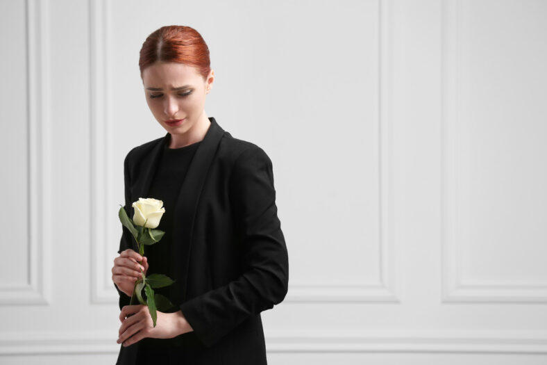Sad woman with rose flower mourning near white wall, space for text. Funeral ceremony