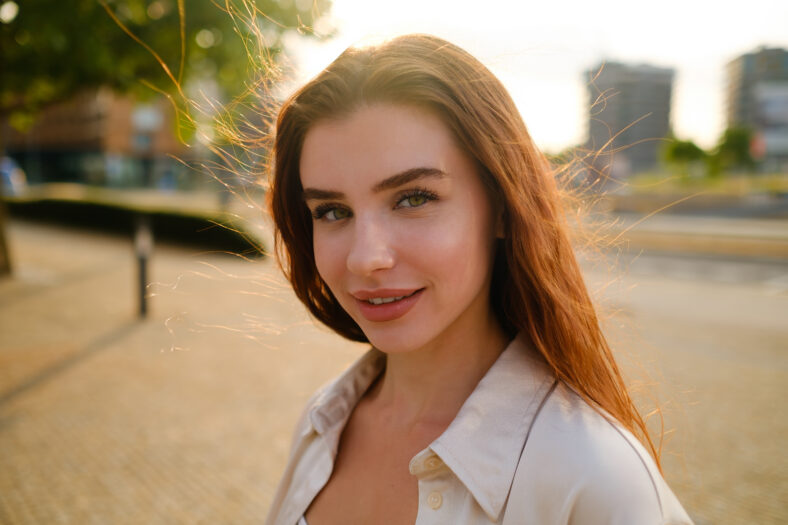 A portrait of a red-haired woman stands on the street, her vibrant locks framing her face.