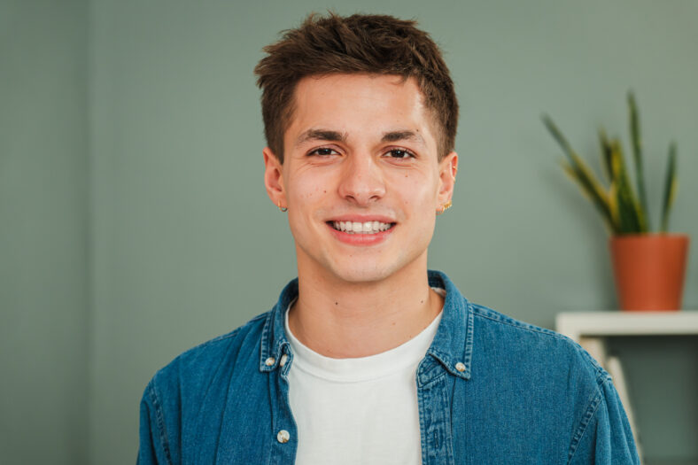 Close up individual portrait of one young adult caucasian guy smiling and looking at camera with friendly expression. Headshot of a real teenage man student with white teeth staring front at home. High quality photo