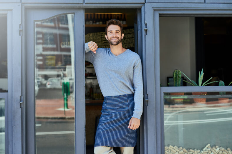 Happy, small business and portrait of man at coffee shop for service industry, welcome or smiling. Confidence, entrepreneur and male barista with apron at cafe entrance, doorway or front door.