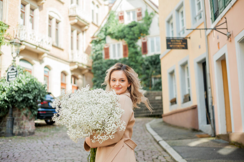 Portrait of beautiful young blonde woman with bouquet of white flowers in the center of European city. Happy model in casual spring clothes.