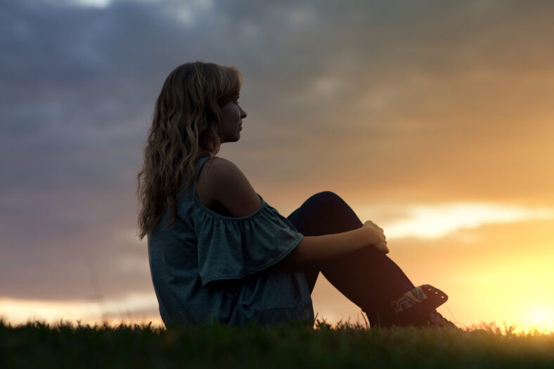 A woman sits in the grass and watches the sunset.
