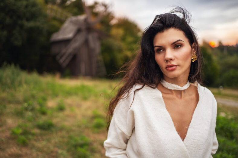 Retro style. Portrait of a young beautiful girl with black chic hair in a white dress on the background of a forest, a mill and a red sunset.