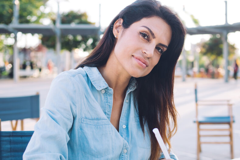 Beautiful caucasian female in casual wear enjoying free time on cafe terrace writing in notepad, charming young 30s woman journalist making notes for creating article blog on sunny day outdoors