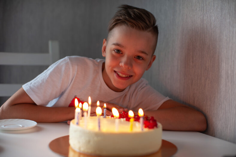 Smiling young boy leaning on a table with a lit birthday cake, enjoying a joyful celebration in a cozy setting.