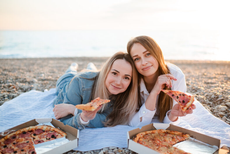 Two girl friends 25-29 year old having picnic with pizza at beach together. Summer vacation season. Looking at camera.