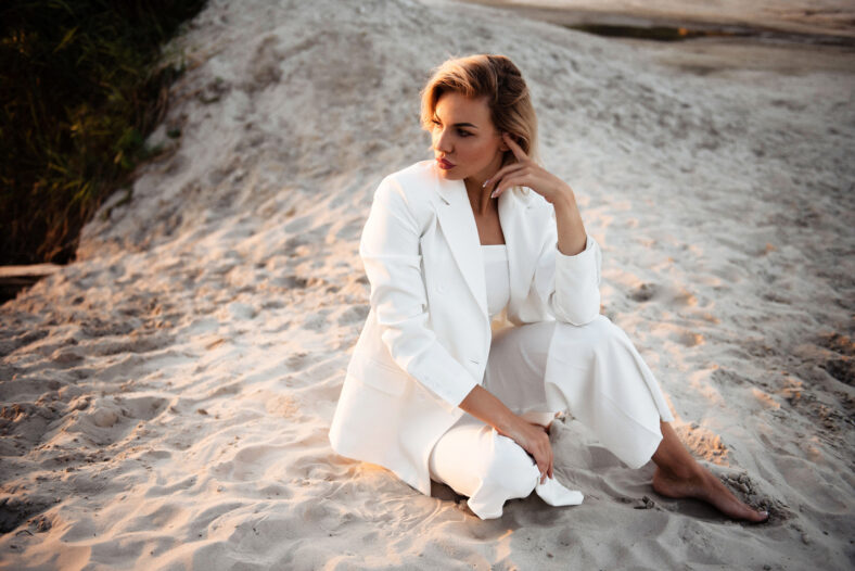 Stunning young woman with blond hair sitting on sand and looking aside during professional photo session on nature. Female model in white suit posing on camera outdoor.