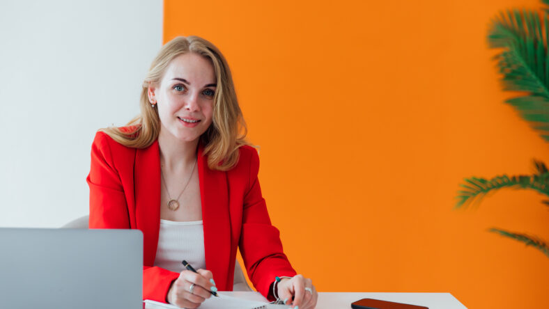 Business blonde woman in red suit at desk with laptop working in office