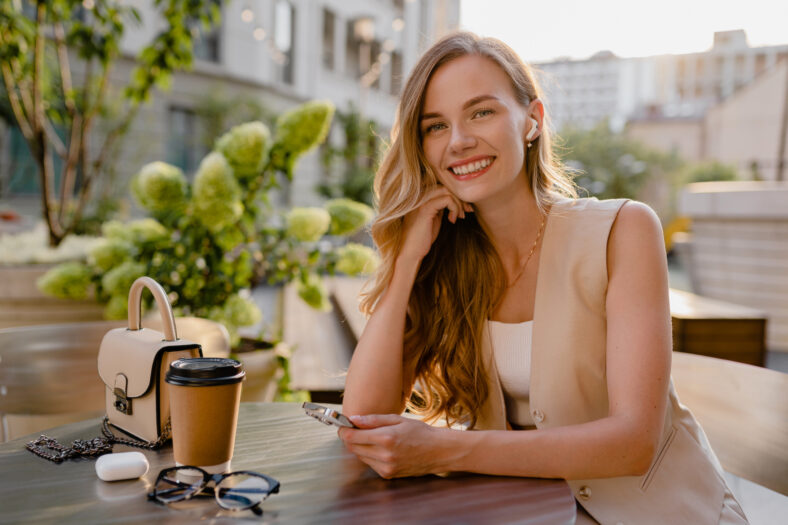 attractive young blond woman sitting in cafe using smart phone drinking coffee in spring summer autumn fashion trend outfit smiling happy wearing beige suit street style, thinking busy and confident