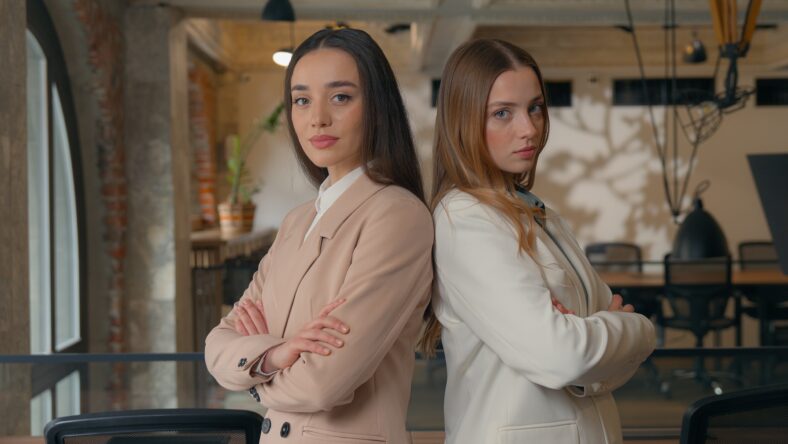 Two confident Caucasian business women girls partners entrepreneurs female businesswomen corporate workers colleagues posing in office crossed hands leaning backs together looking at camera teamwork. High quality 4k footage