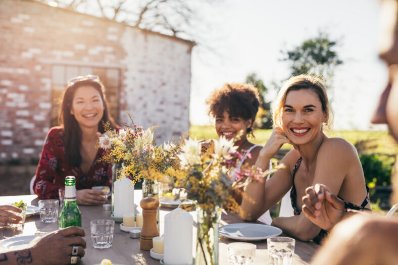 Young people partying in a restaurant outdoors. Multiracial friends sitting together around a table and smiling during garden party.