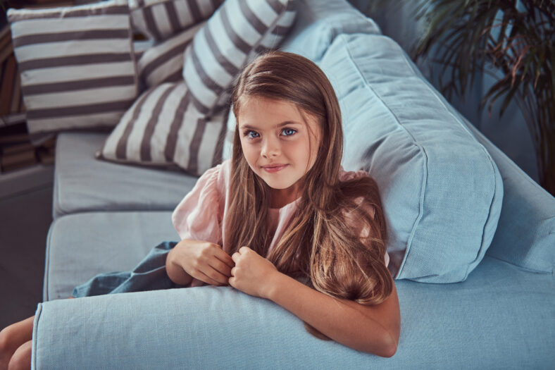 Portrait of a smiling little girl with long brown hair and piercing glance, lying on a sofa at home.