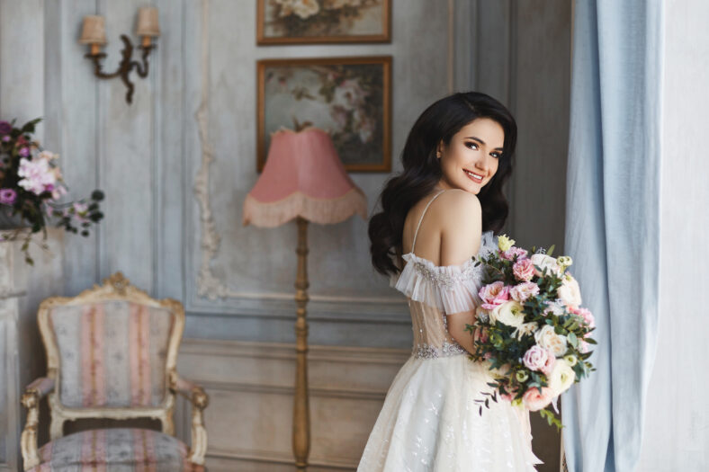 Beautiful fashionable bride, young model brunette woman in stylish wedding dress with naked shoulders with bouquet of flowers in her hands posing in luxury vintage interior near the window.