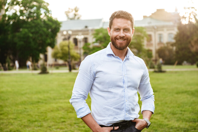 Portrait of european attractive man in white shirt standing with hands in pockets during walk in green park over old building background