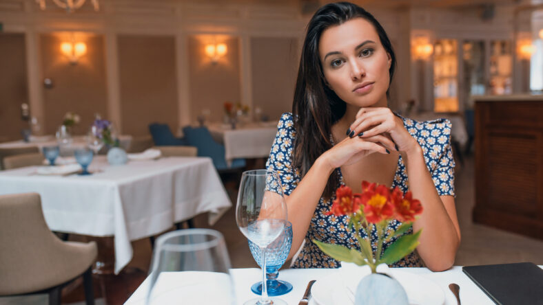 Horizontal indoor portrait of brunette young elegant woman wearing dress sitting in the restaurant looking to the camera. Caucasian female posing in cafeteria and waiting her friends. Copy space