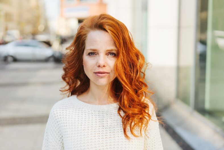 Serious attractive young woman with long red hair looking pensively at the camera as she stands in a city street