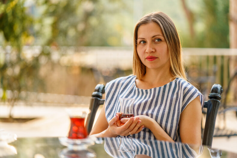 Beautiful young woman with a dreamy lip smile on her face is having cup of tea while sitting at the outdoor park terrace at the mirror surface table. Green nature surrounding. Summer vacation day.