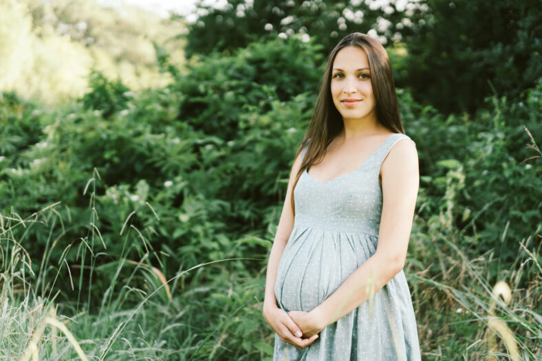 Beautiful caucasian pregnant brunette woman with long hair in a delicate dress in the park hugs her tummy.