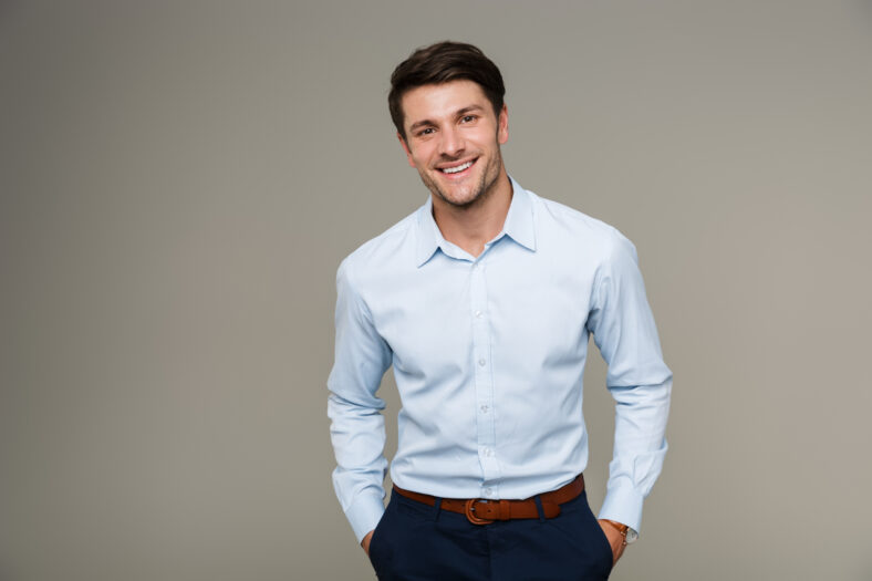 Image of happy brunette man wearing formal clothes smiling at camera with hands in pockets isolated over gray background