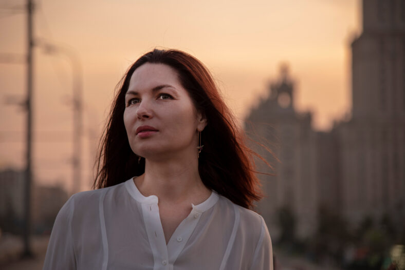 Portrait of a middle-aged woman in profile in a white blouse with her hair down. Against the background of the evening city. A dreamy look into the distance. Houses, sunset.