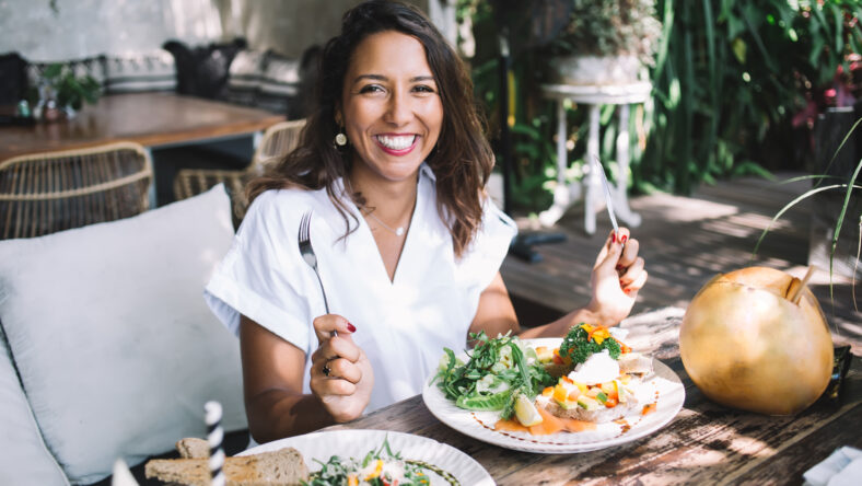 Happy ethnic lady with fork and knife sitting at wooden table with local exotic dishes and drinks in comfortable outdoor cafe on blurred background