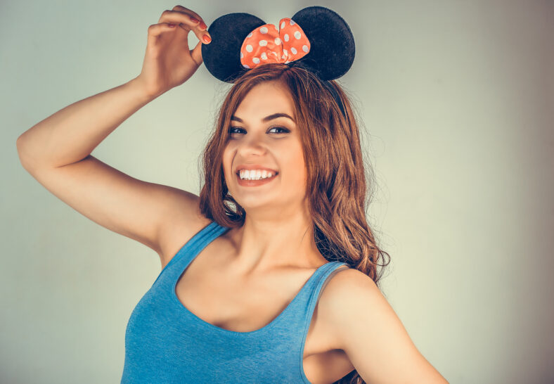 Closeup portrait of young woman wearing blue shirt and black, red with white polka dots mouse ears playing flirty. Mixed race model isolated on light green copy space background. Horizontal image
